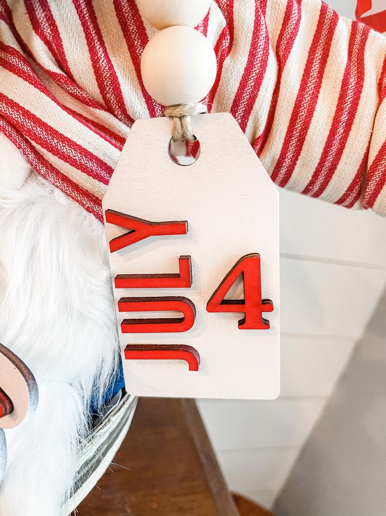 Puede incluir: Una etiqueta de madera blanca con letras rojas que dicen "July 4" colgando de una tela a rayas rojas y blancas. La etiqueta est&aacute; unida a una cuerda de cuentas de madera.