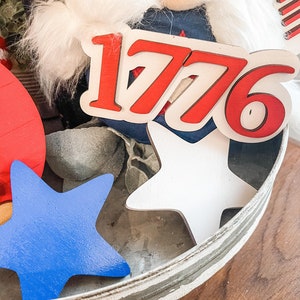 Puede incluir: Una bandeja blanca de varios niveles con decoraciones rojas, blancas y azules. La bandeja presenta un letrero de madera con el n&uacute;mero "1776" en letras rojas, dos estrellas blancas y una estrella azul.