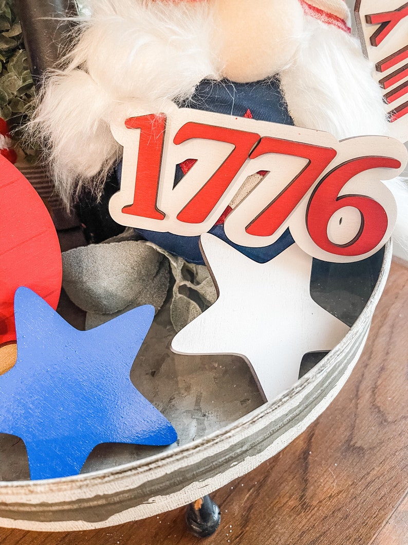 Puede incluir: Una bandeja blanca de varios niveles con decoraciones rojas, blancas y azules. La bandeja presenta un letrero de madera con el n&uacute;mero "1776" en letras rojas, dos estrellas blancas y una estrella azul.