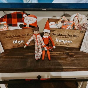 Op de afbeelding: Twee houten kerstavonddozen met de namen Mya en Harper, gevuld met kerstartikelen zoals warme chocolademelk en knuffels. Twee elfpoppen zitten voor de dozen, geflankeerd door twee geprinte brieven gericht aan Mya en Harper.