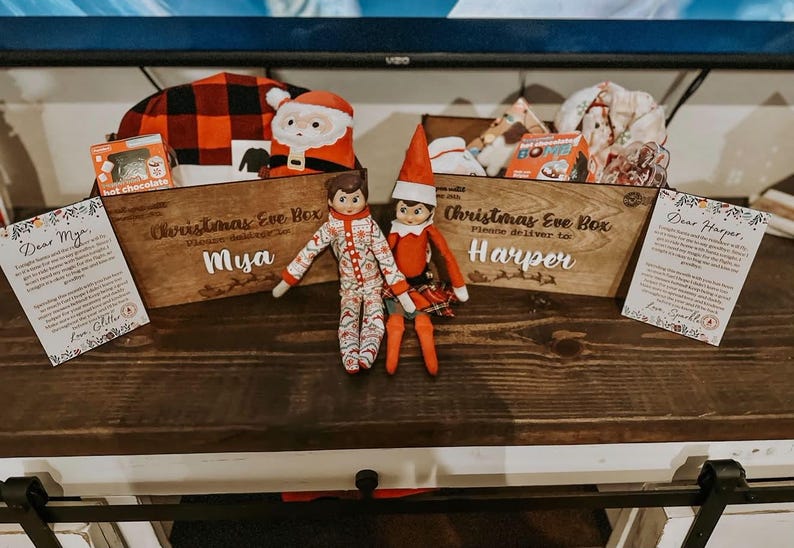 Op de afbeelding: Twee houten kerstavonddozen met de namen Mya en Harper, gevuld met kerstartikelen zoals warme chocolademelk en knuffels. Twee elfpoppen zitten voor de dozen, geflankeerd door twee geprinte brieven gericht aan Mya en Harper.
