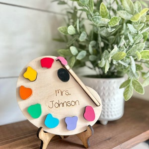 May include: Wooden paint palette with colourful paint swatches and a paintbrush. The palette has the name "Mrs. Johnson" written on it.