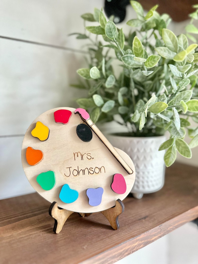 May include: Wooden paint palette with colourful paint swatches and a paintbrush. The palette has the name "Mrs. Johnson" written on it.