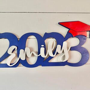 Grad Sign shown with 2023 with the 2023 in Royal Blue, The Hat in Red and the name in white by walnut + birch