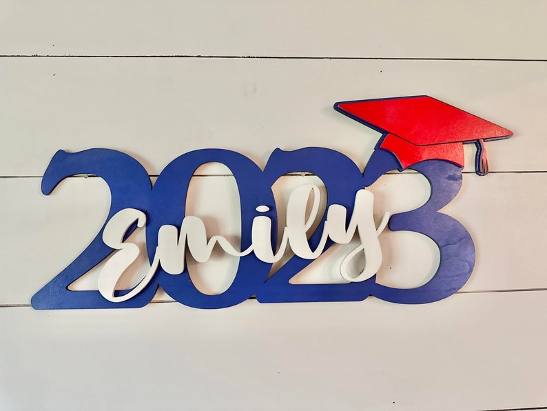 Grad Sign shown with 2023 with the 2023 in Royal Blue, The Hat in Red and the name in white by walnut + birch