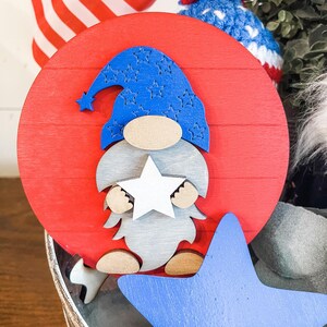 Puede incluir: Un c&iacute;rculo de madera rojo con una estrella blanca y un gnomo azul sosteniendo una estrella blanca. El gnomo lleva un sombrero azul con estrellas blancas. El c&iacute;rculo est&aacute; sentado sobre una superficie de madera con una estrella azul.