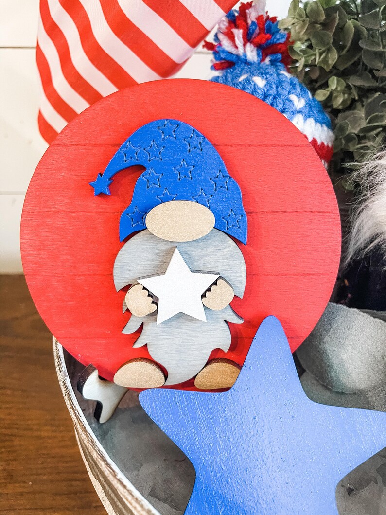 Puede incluir: Un c&iacute;rculo de madera rojo con una estrella blanca y un gnomo azul sosteniendo una estrella blanca. El gnomo lleva un sombrero azul con estrellas blancas. El c&iacute;rculo est&aacute; sentado sobre una superficie de madera con una estrella azul.