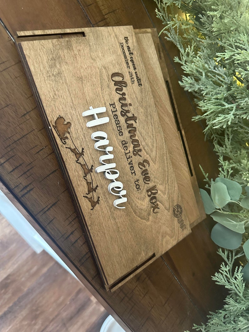 Op de afbeelding: Een rechthoekige houten doos met een lasergegraveerd ontwerp van een arrenslee met rendieren en de tekst: Do not open until DECEMBER 24th, Christmas Eve Box, Please deliver to: Harper. De naam Harper is in verhoogde witte letters.