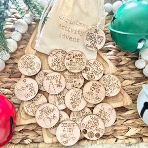 May include: A set of 17 wooden Christmas activity tokens with various holiday-themed activities written on them, such as "Listen to Holiday Music", "Bake Some Christmas Cookies", "Decorate the Tree", and "Play a Game with Family". The tokens are arranged on a woven basket with a white drawstring bag and two jingle bells.