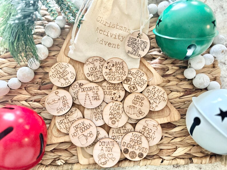 May include: A set of 17 wooden Christmas activity tokens with various holiday-themed activities written on them, such as "Listen to Holiday Music", "Bake Some Christmas Cookies", "Decorate the Tree", and "Play a Game with Family". The tokens are arranged on a woven basket with a white drawstring bag and two jingle bells.