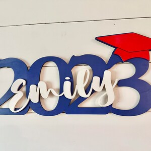 Grad Sign shown with 2023 with the 2023 in Royal Blue, The Hat in Red and the name in white by walnut + birch