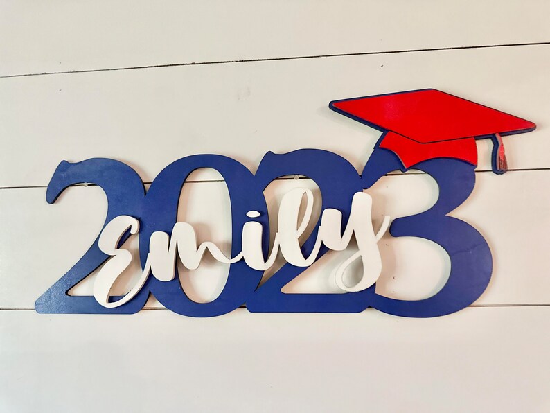 Grad Sign shown with 2023 with the 2023 in Royal Blue, The Hat in Red and the name in white by walnut + birch