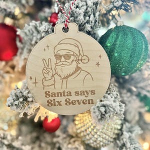 May include: Wooden Christmas ornament with a laser-etched design of Santa Claus wearing sunglasses and giving a peace sign. The text "Santa says Six Seven" is below the image. The ornament hangs from a red and white string.