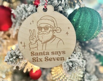 May include: Wooden Christmas ornament with a laser-etched design of Santa Claus wearing sunglasses and giving a peace sign. The text "Santa says Six Seven" is below the image. The ornament hangs from a red and white string.