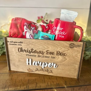 A wooden box with the text "Christmas Eve Box" and "Please deliver to: Harper". The box contains a red mug with the text "Hot Cocoa 50¢", a package of hot chocolate mix, and a red and white blanket with a unicorn design. Box ONLY for sale.