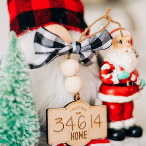 May include: A wooden Christmas ornament with a red and black buffalo plaid gnome, a white bow, and a wooden sign that reads "Christmas 2021 34614 Home".