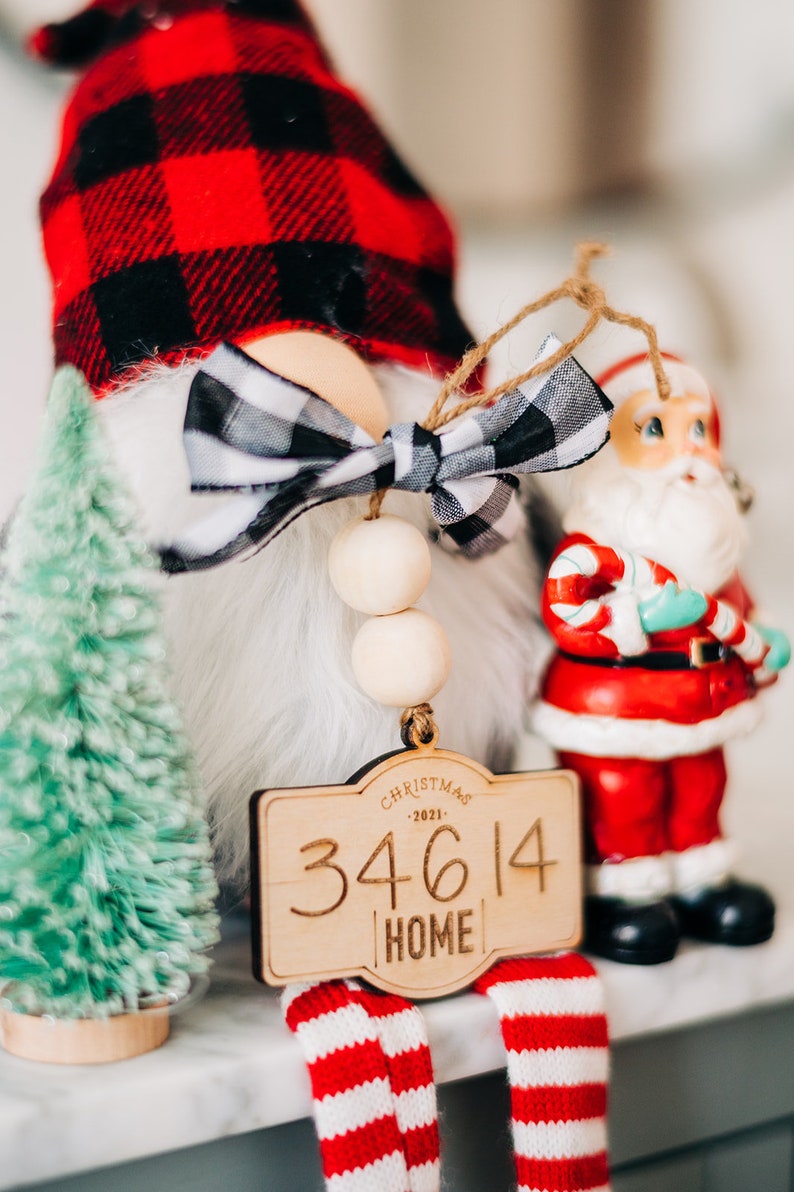 May include: A wooden Christmas ornament with a red and black buffalo plaid gnome, a white bow, and a wooden sign that reads "Christmas 2021 34614 Home".