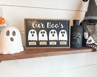 May include: A wooden sign with the text "Our Boo's" and four white ghost cutouts with names below each ghost: Justin, Kaylee, Mya, and Harper. The sign is on a wooden shelf with a black bottle that says "October 31" and a black and white gnome.