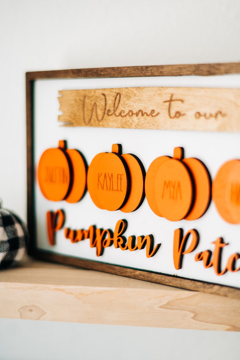 May include: A wooden sign with the text "Welcome to our Pumpkin Patch" in orange lettering. There are five orange pumpkin-shaped ornaments with names written on them.