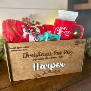 Op de afbeelding: Een houten kerstavonddoos met de tekst Do not open until December 24th, Christmas Eve Box, Please deliver to: Harper en een arrenslee-afbeelding. Binnenin een rode mok met Hot Cocoa 50&cent;, Ghirardelli-chocolade en een rode eenhoorndeken.