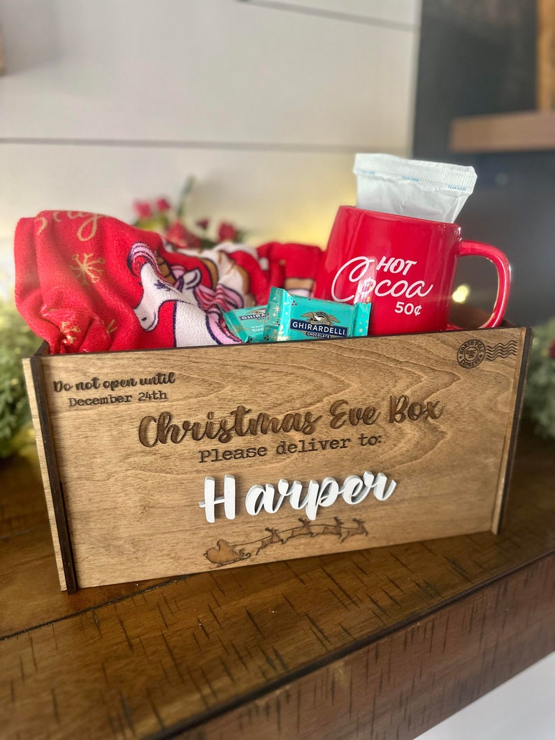 Op de afbeelding: Een houten kerstavonddoos met de tekst Do not open until December 24th, Christmas Eve Box, Please deliver to: Harper en een arrenslee-afbeelding. Binnenin een rode mok met Hot Cocoa 50&cent;, Ghirardelli-chocolade en een rode eenhoorndeken.