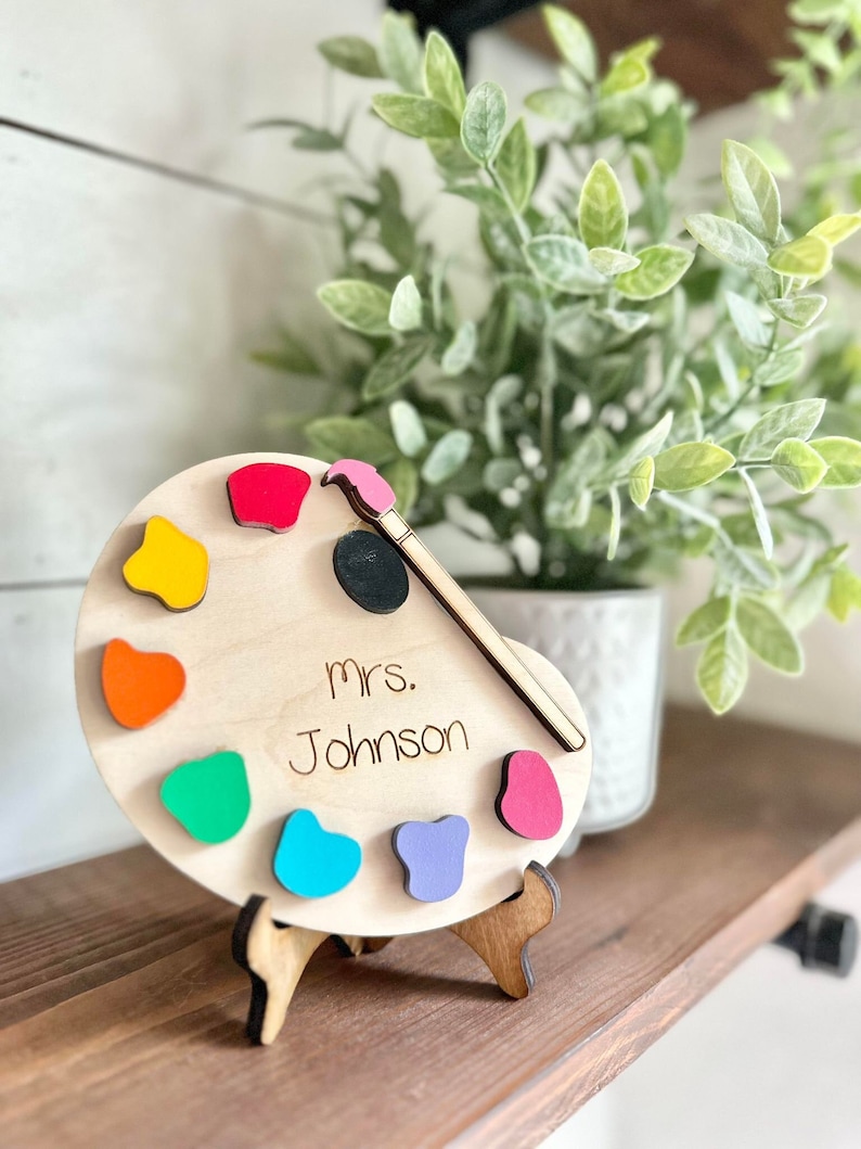 May include: Wooden paint palette with a paintbrush and colorful paint swatches. The palette has the name "mrs. Johnson" written on it.
