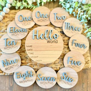 May include: A set of wooden baby milestone discs. The central disc says "Hello World" and has space for baby's information. Surrounding discs mark months one through eleven, plus "One Year". The text is light blue.