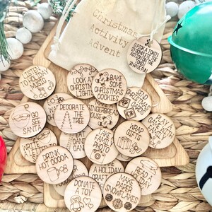 May include: A set of 16 wooden Christmas activity tokens with various holiday-themed illustrations and text. The tokens are in a small burlap bag with the words "Christmas Activity Advent" printed on it.  The tokens include activities such as "Listen to Holiday Music", "Go See Santa", "Write a Letter to Santa", "Drink Hot Cocoa", "Decorate the Tree", "Bake Some Christmas Cookies", "Make Gingerbread House", "Take Family Photo", "Wrap Gifts", "Send Christmas Cards", "Play a Game with Family", "Read a Holiday Book", "Make Paper Snowflakes", "Donate a Toy", "Eat Candy Cane", "A Holiday Craft", and "Look at Christmas Lights".