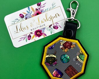 May include: A black and gold octagonal keychain featuring embroidered dice and class symbols. The keychain has a silver clasp and a black leather backing. A business card with floral accents and the text "Lilies & Larkspur Boutique" is also visible.