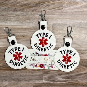 May include: Three white round keychains with black stitching and a red medical symbol with a black outline. Each keychain has the text "TYPE I DIABETIC" in black.
