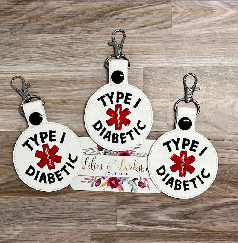 May include: Three white round keychains with black stitching and a red medical symbol with a black outline. Each keychain has the text "TYPE I DIABETIC" in black.