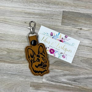 May include: A brown leather keychain featuring a German Shepherd dog design with a silver-colored clasp. The keychain is next to a white business card with the text "Lilies & Larkspur Boutique" and floral accents.
