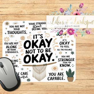 May include: White mouse pad with a motivational quote: "It's okay not to be okay." The quote is surrounded by stars, flowers, and other decorative elements. The text "You are not your thoughts" and "You are capable" are also included.