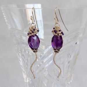 May include: A pair of silver earrings with amethyst gemstones and wire wrapped details. The earrings feature a dangling amethyst gemstone and a silver wire that is twisted into a spiral shape.