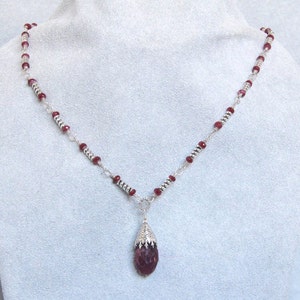 Puede incluir: Un collar de cadena de plata con un colgante en forma de lágrima que presenta una piedra preciosa de color rojo intenso. La cadena está adornada con pequeñas cuentas de plata y cuentas rojas.