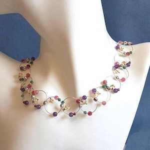 Op de afbeelding: Een ketting bestaande uit een reeks onderling verbonden dunne zilverkleurige metalen ringen. Het ontwerp is geaccentueerd met kleine, ronde kralen in diverse kleuren, waaronder paars, groen, roze en transparant, verspreid over de metalen schakels.