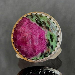 May include: A round ring featuring a large, polished cabochon stone with a vibrant magenta-pink center and mottled green and black patterns. The stone is set within a metallic bezel with a decorative, scalloped edge along the outer rim.