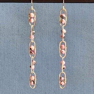 May include: A pair of dangle earrings with a chain design. Each earring features a series of oval-shaped links connected by small pink and black beads. The earrings are suspended from a simple hook.