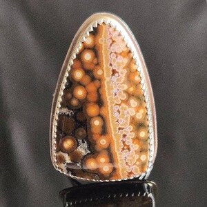 May include: A teardrop-shaped gemstone ring with a silver bezel. The stone features a unique pattern of brown, orange, and white circular spots. The ring's edge has a decorative, serrated design.