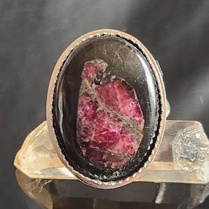 Puede incluir: Anillo ovalado de plata con una piedra oscura grande y una gema rojo-rosa texturizada en el centro. El engaste del anillo es de plata pulida y la gema tiene un aspecto natural único. El anillo se exhibe sobre un cristal claro y facetado.