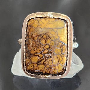 Może przedstawiać: Prostokątny pierścionek z dużym, złoto-brązowym kamieniem o skomplikowanych, pajęczynowych wzorach. Kamień osadzony jest w srebrnej metalowej ramie z teksturowaną krawędzią. Obręcz pierścionka jest również srebrna.