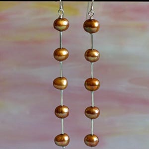 May include: A pair of dangle earrings featuring five bronze-colored, round beads strung on a silver-colored wire. The earrings hang from a silver-colored hook. The background is a soft blend of pink and white.