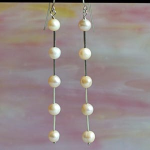 May include: Elegant dangle earrings featuring multiple creamy white pearls strung on delicate silver-tone wires. The earrings hang from simple silver hooks, creating a graceful, flowing design. The pearls are evenly spaced, adding a touch of classic sophistication to the jewelry.