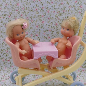 May include: A yellow double stroller with two pink seats and a pink table in the middle. Two baby dolls are sitting in the seats.