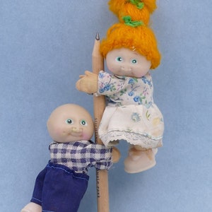 Puede incluir: Dos pequeñas muñecas, una calva y otra con pelo de hilo naranja, están colocadas en un lápiz. La muñeca calva lleva una camisa a cuadros azul y blanco y pantalones azules. La otra muñeca lleva una blusa floral y una falda blanca. El lápiz tiene texto.
