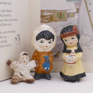 Könnte beinhalten: Drei kleine Keramikfiguren von Kindern in Winterkleidung. Die Figur links trägt einen weißen Mantel und Hut. Die Figur in der Mitte trägt einen braunen Mantel und Hut mit einem blauen Schal. Die Figur rechts trägt ein gelbes Kleid und einen braunen Hut mit einer Feder.