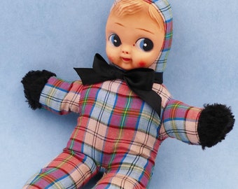 Puede incluir: Un muñeco de peluche con un cuerpo de tela a cuadros y detalles de piel negra. El muñeco tiene una cabeza grande con una cara rosa, ojos azules y una pajarita negra.