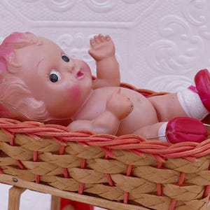 May include: A vintage baby doll with pink hair and rosy cheeks lies in a woven basket. The doll wears red shoes and white socks. The basket is made of light brown wicker with a red trim.