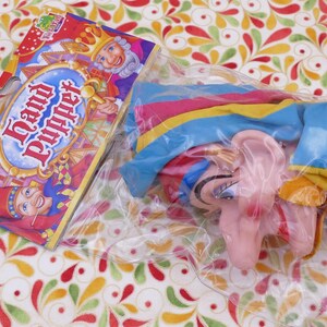 Puede incluir: Un títere de mano rosa con un sombrero azul, amarillo y rosa. El títere tiene una nariz grande y una gran sonrisa. El títere está empaquetado en una bolsa de plástico transparente con una etiqueta colorida que dice "Títere de mano".