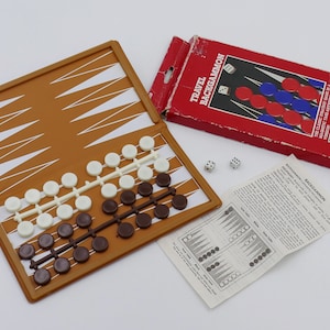 Puede incluir: Un juego de backgammon de viaje de color marrón con un tablero plegable, fichas blancas y marrones y dos dados. La caja es roja con las palabras "Travel Backgammon" impresas en ella.